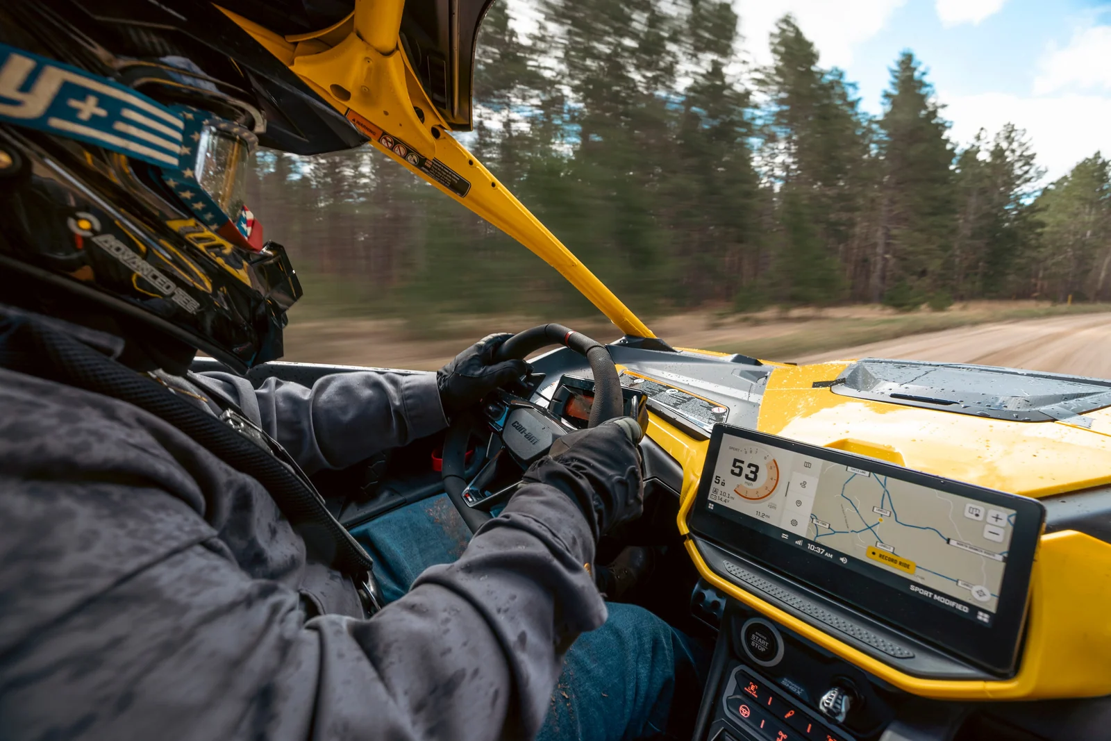 A helmeted driver steers a Can-Am Maverick R along a forest trail, viewed from inside the cockpit.
