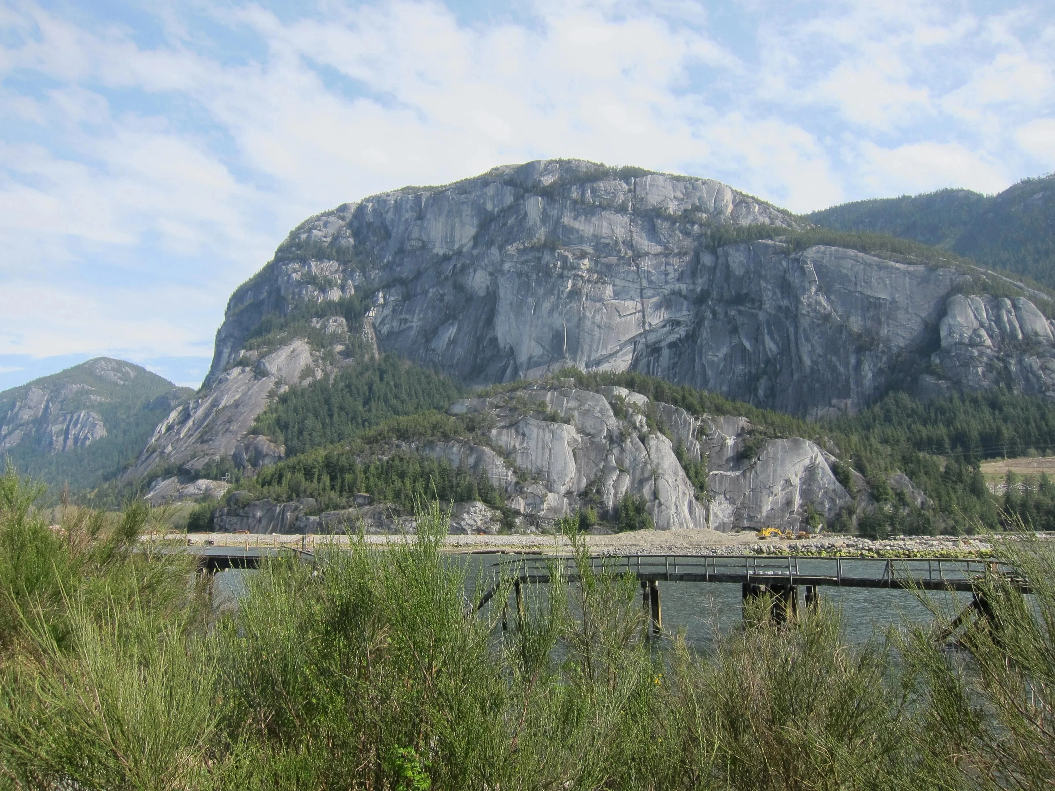 A broad granite cliff rises above trees, a narrow waterfront and a wooden bridge.