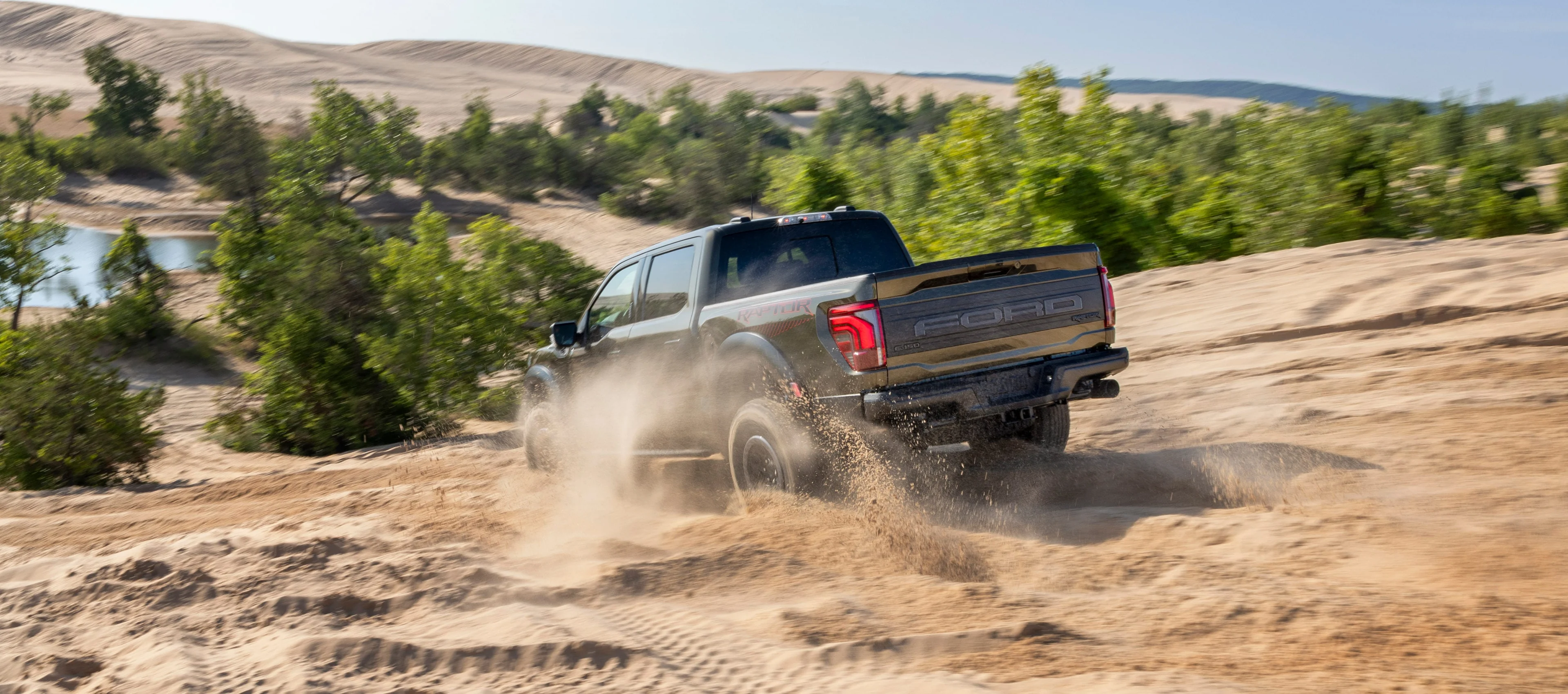 A 2024 Ford F-150 Raptor drives away up a broad sandy slope, with wheel tracks leading through the dunes.