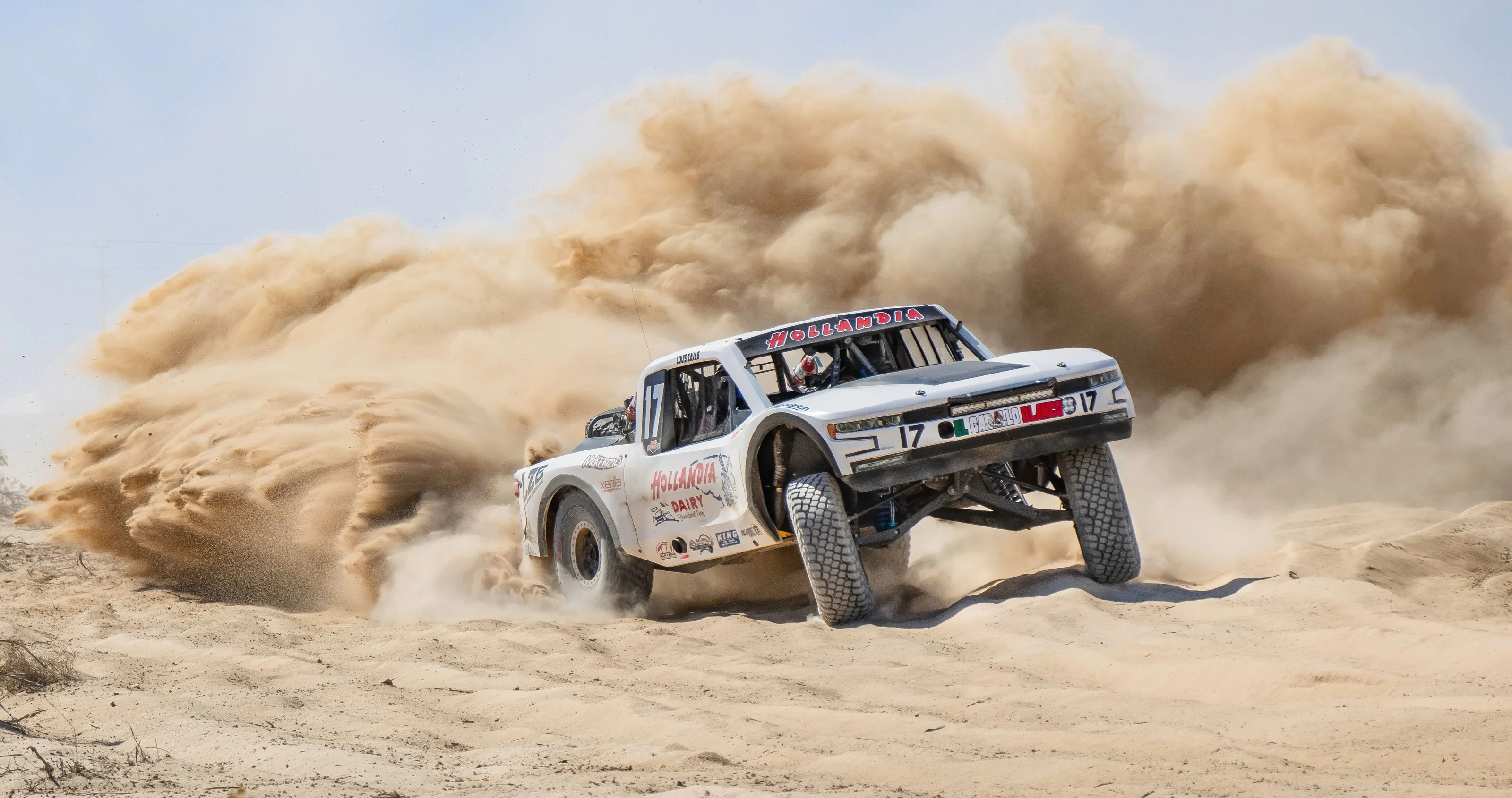 A race truck crosses rough desert ground at speed, with helmeted crew visible.