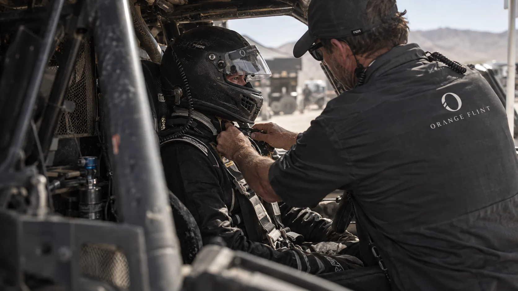 An Orange Flint technician adjusts a helmeted driver’s harness inside a race cockpit.