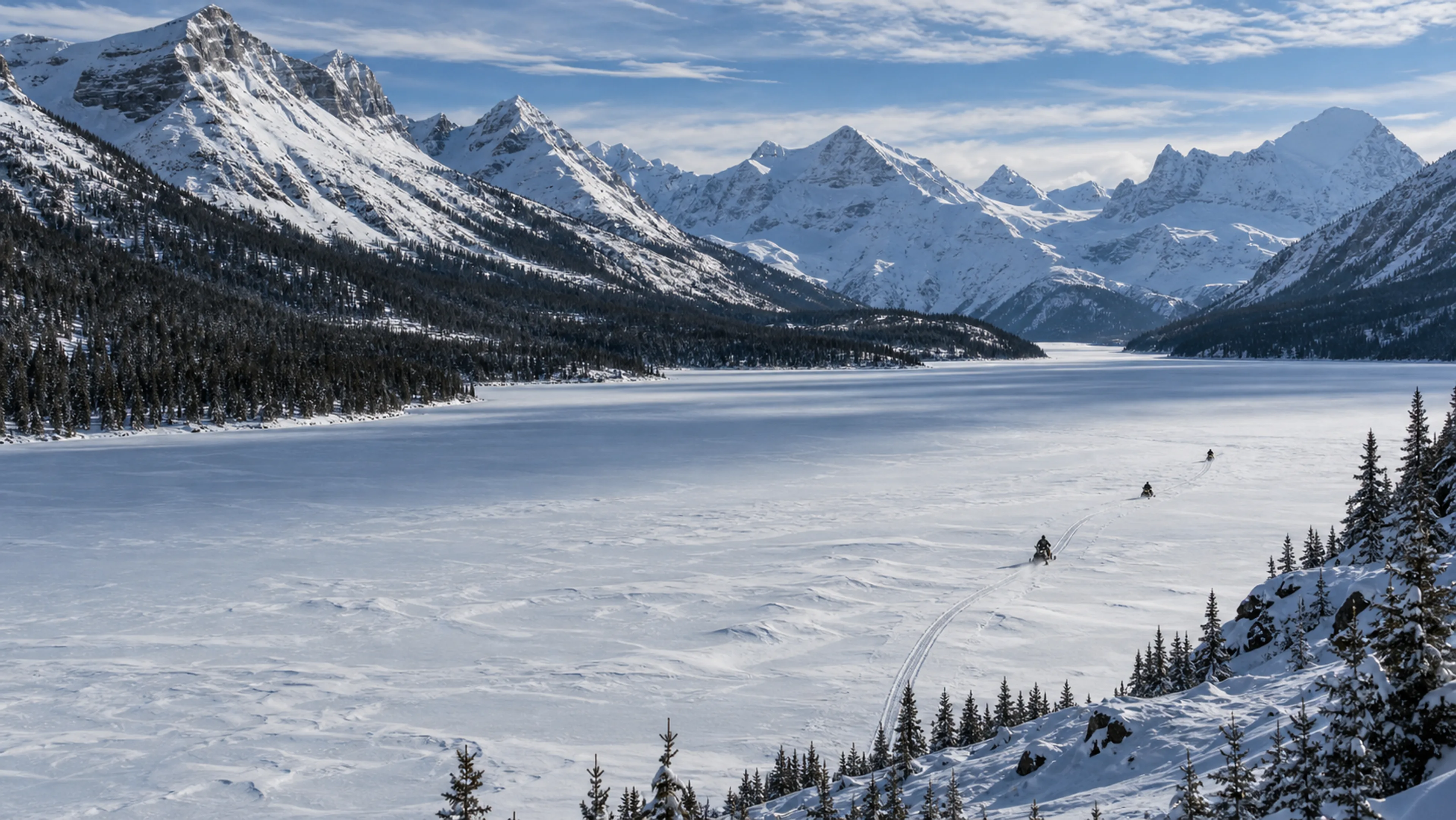 Three small snowmobiles follow a track across an immense frozen mountain lake.
