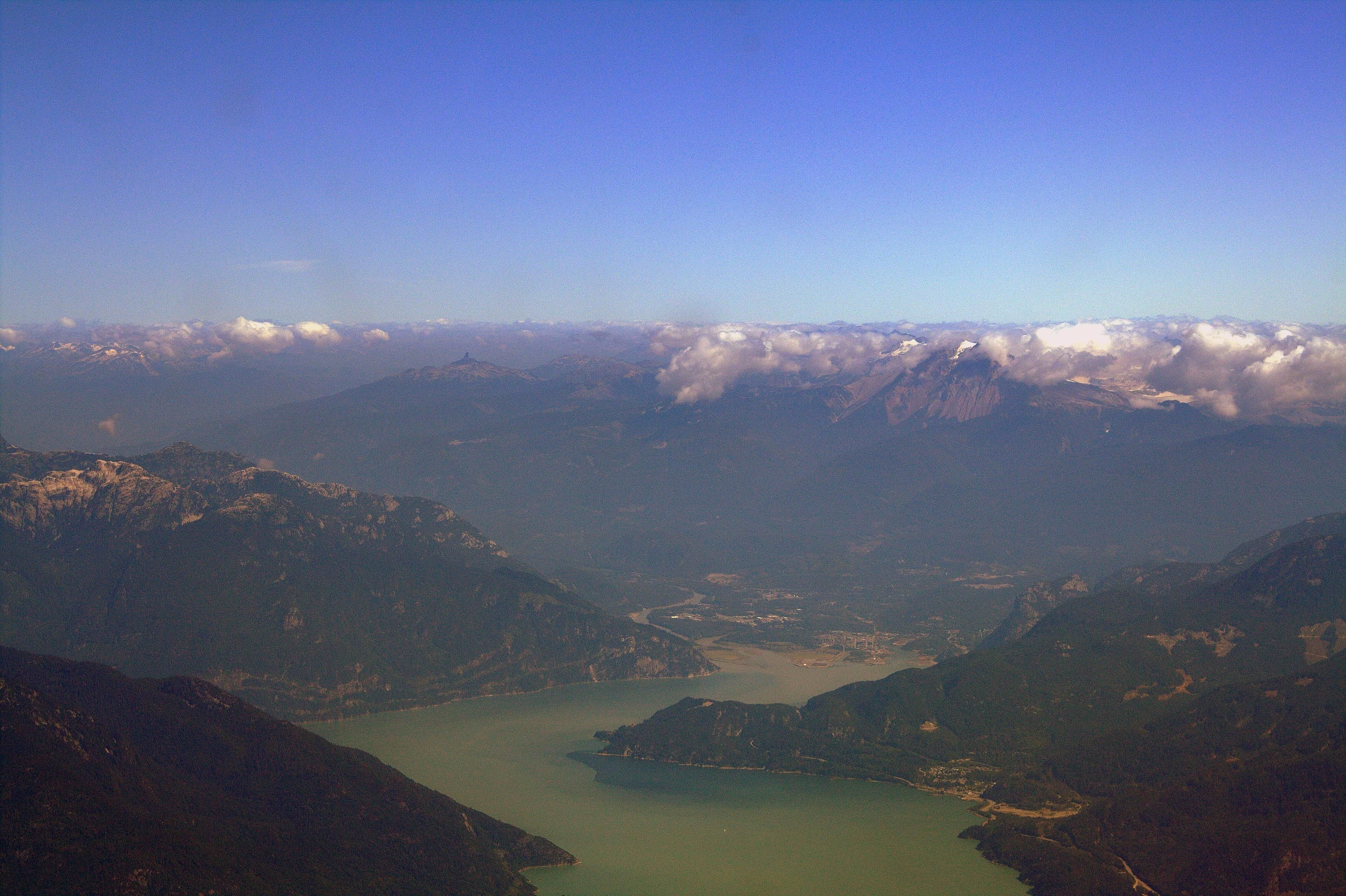 Aerial view of an inlet narrowing between forested mountains beneath a cloud-fringed horizon.