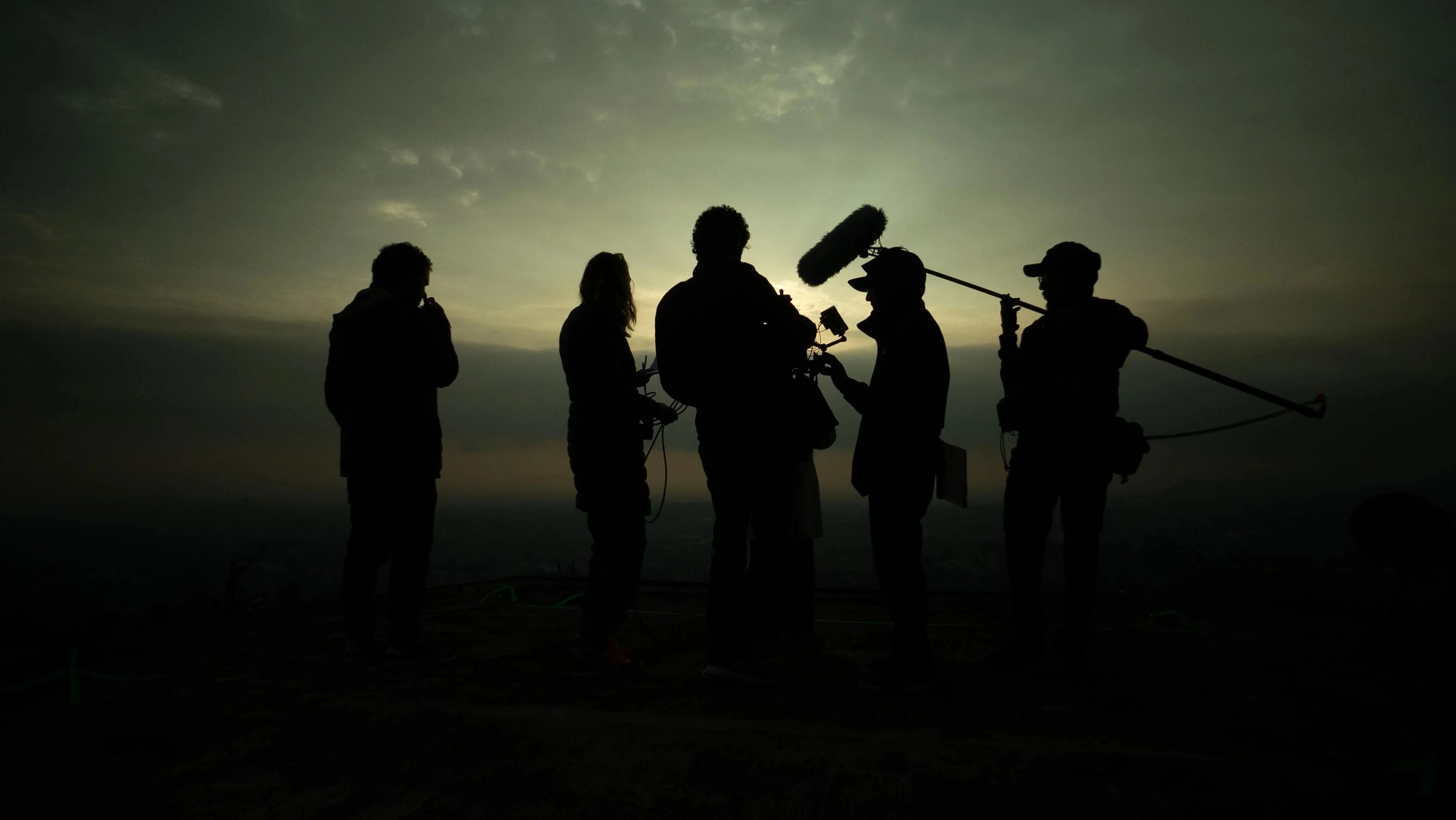 An outdoor film crew works with a camera and boom microphone, silhouetted against the evening sky.