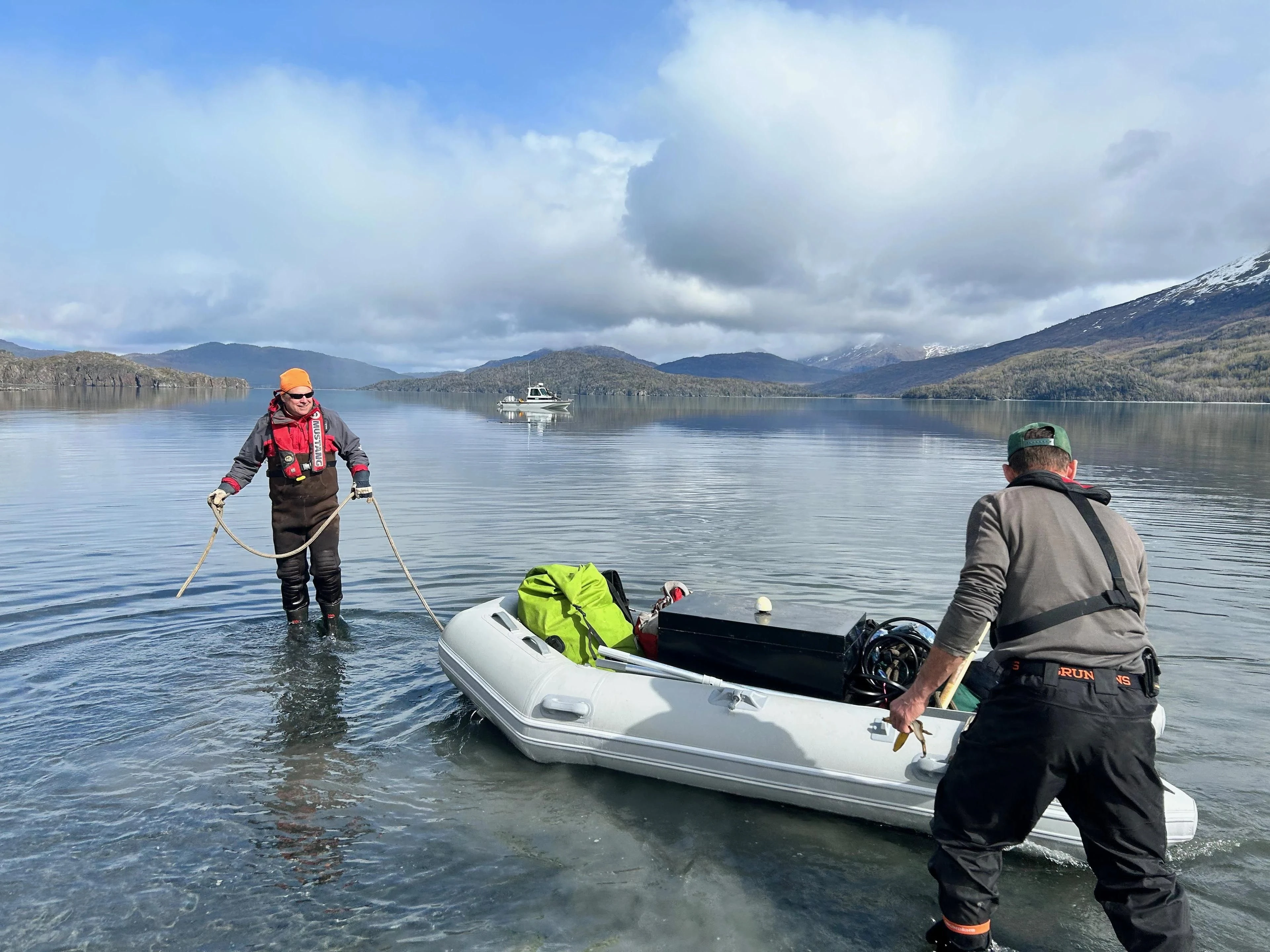 Two field researchers bring an equipment-laden inflatable boat ashore while a support boat waits offshore.