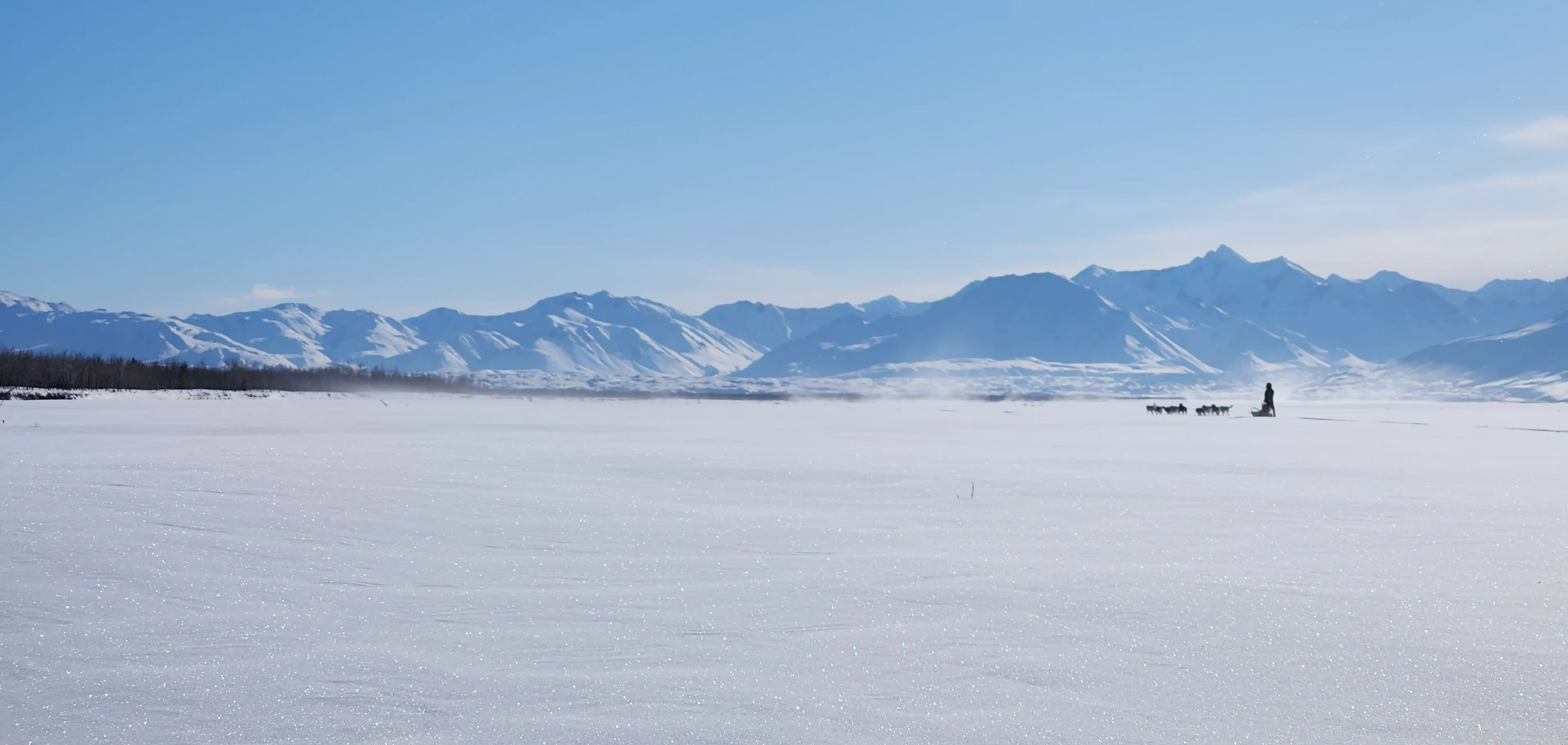 A distant dog team crosses a broad snow-covered river basin below an open mountain horizon.