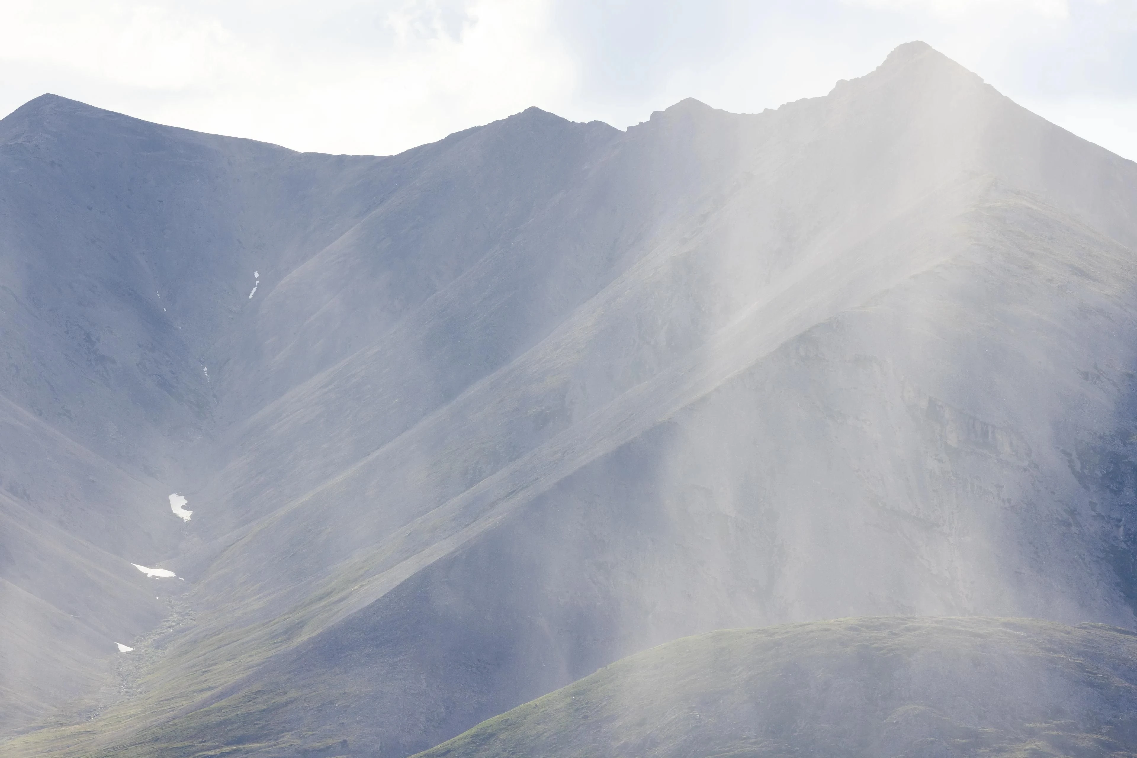 A pale curtain of rain partially obscures a steep mountain slope, with small snow patches in a gully.