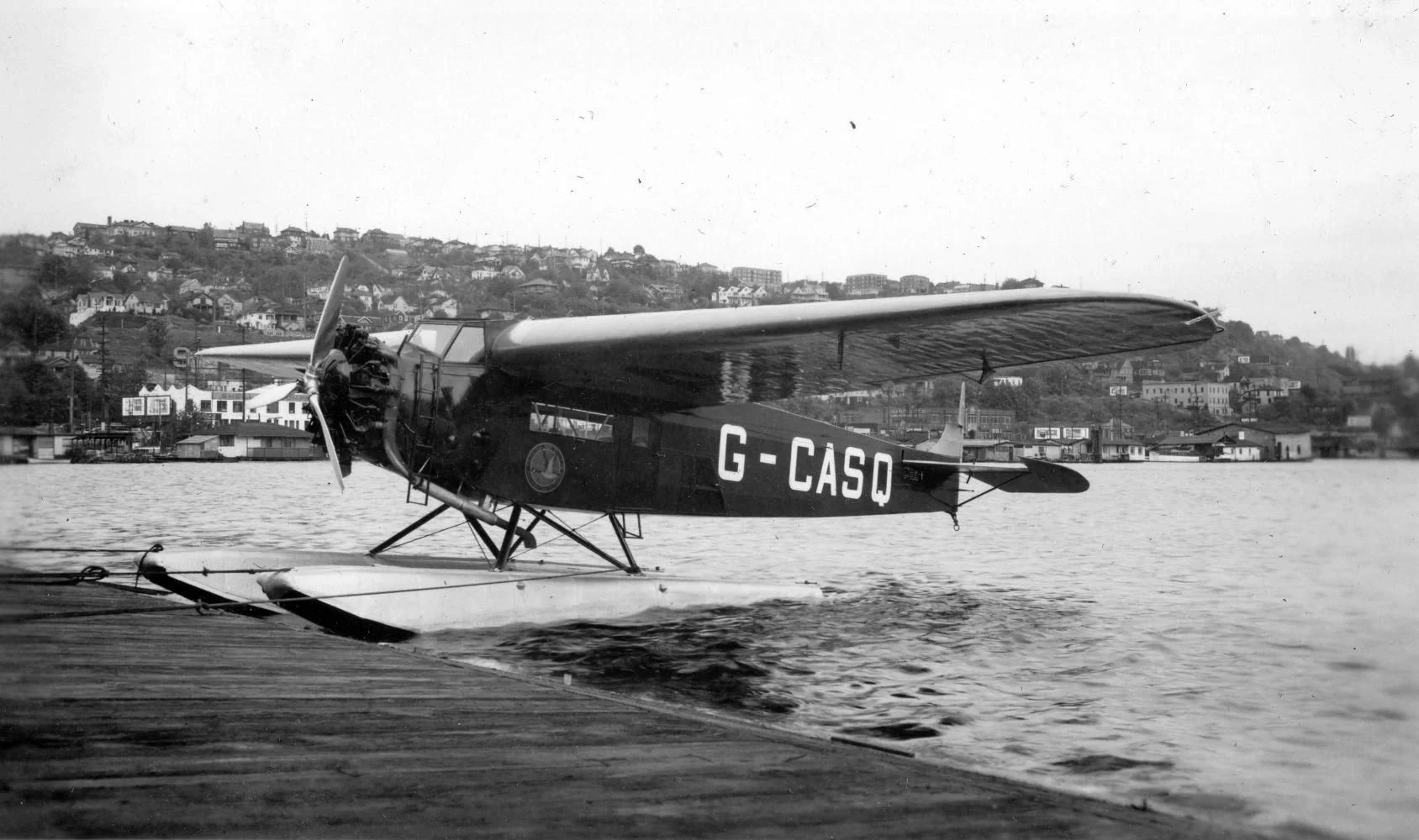 A Fokker Super Universal marked G-CASQ rests on twin floats beside a wooden dock, with a hillside town across the water.