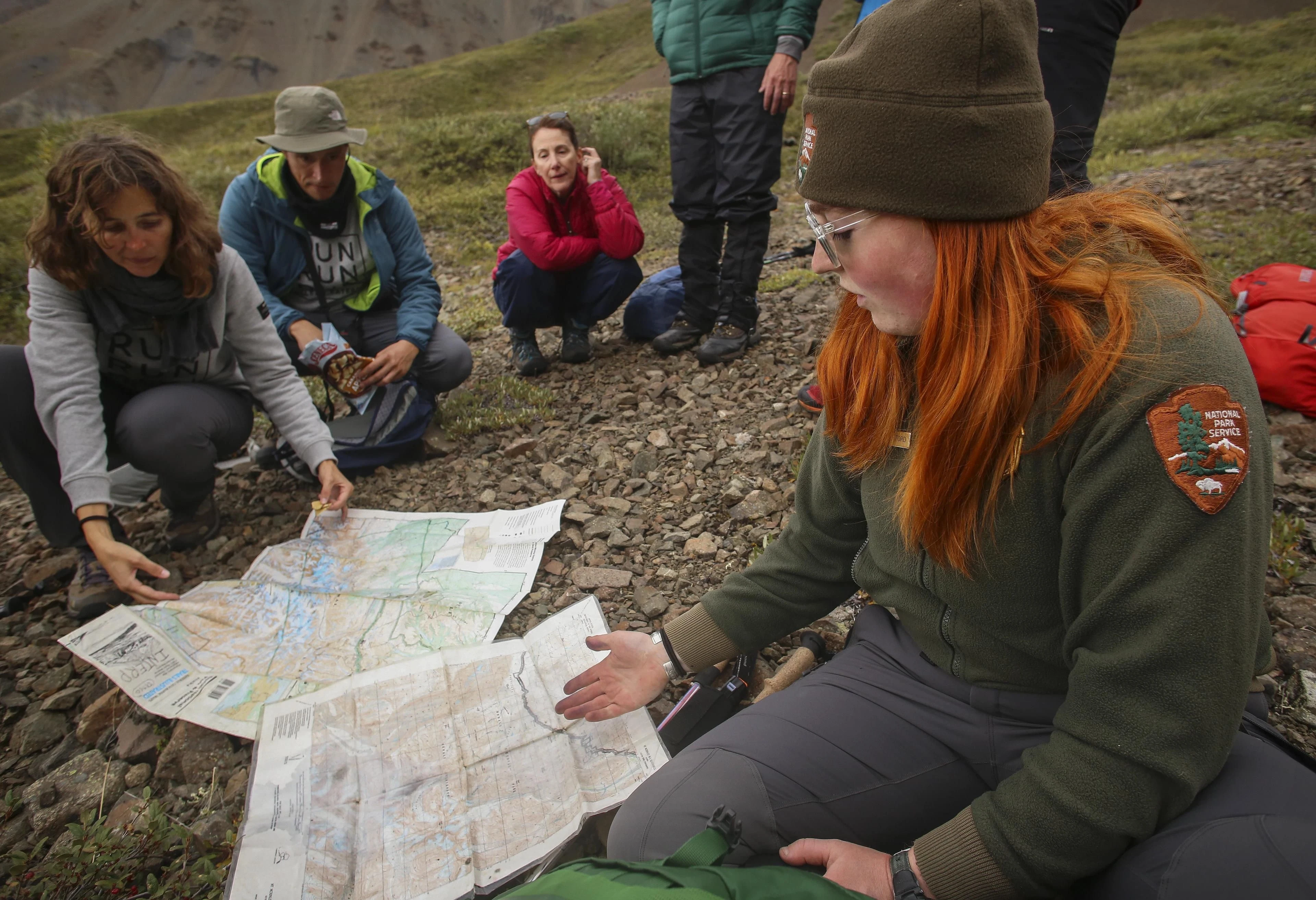 A ranger explains a topographic map to hikers seated on an open hillside.