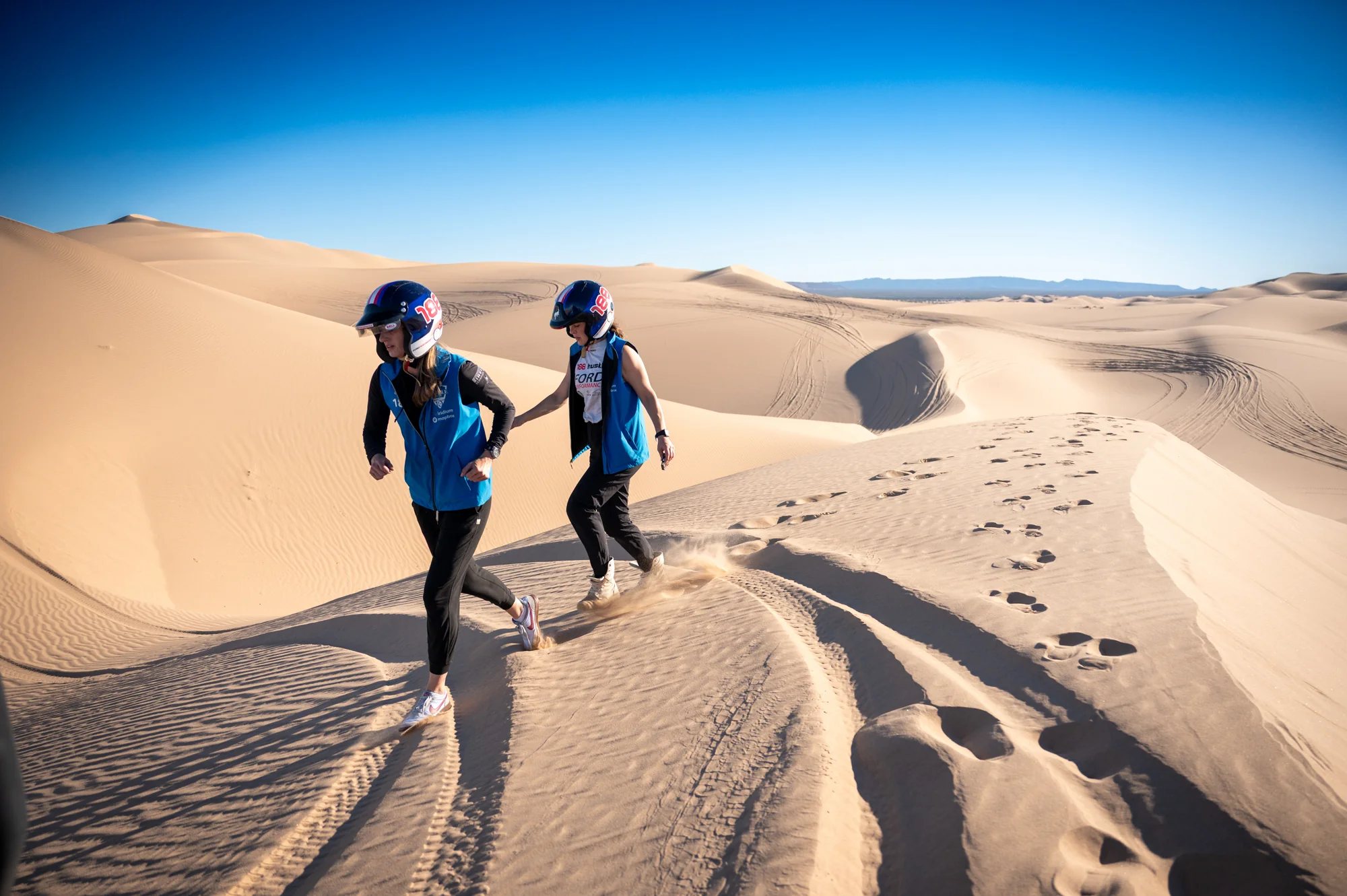 Two helmeted Rebelle Rally competitors walk along a sand ridge, leaving footprints beside the vehicle tracks.