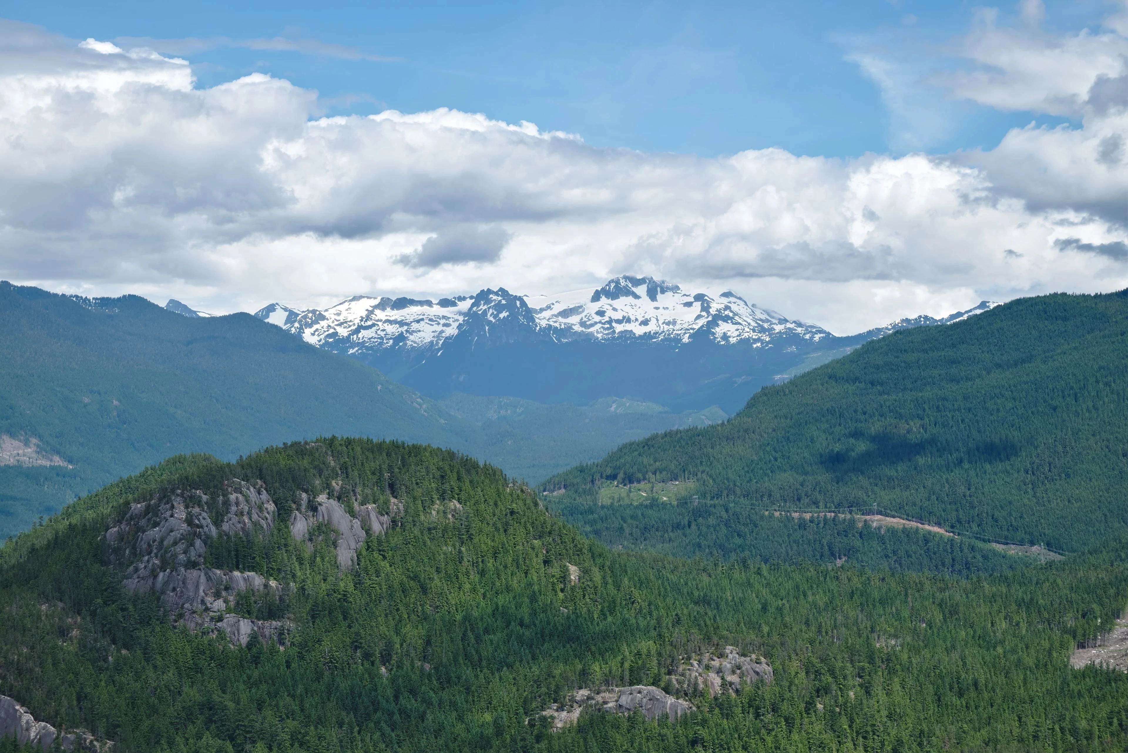 Forested hills and exposed granite sit below distant snow-covered mountains and broken cloud.