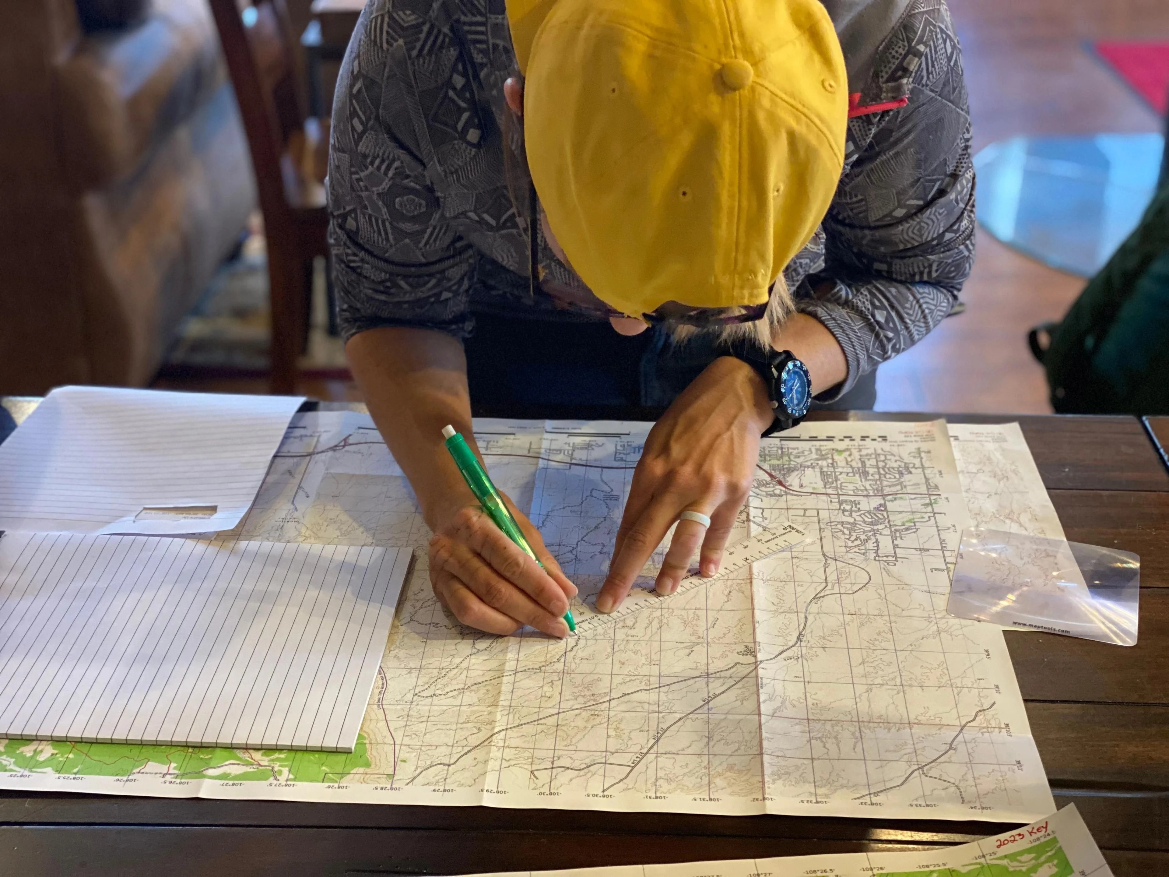 Hands mark a route on a folded topographic map beside an open notebook and other working maps.