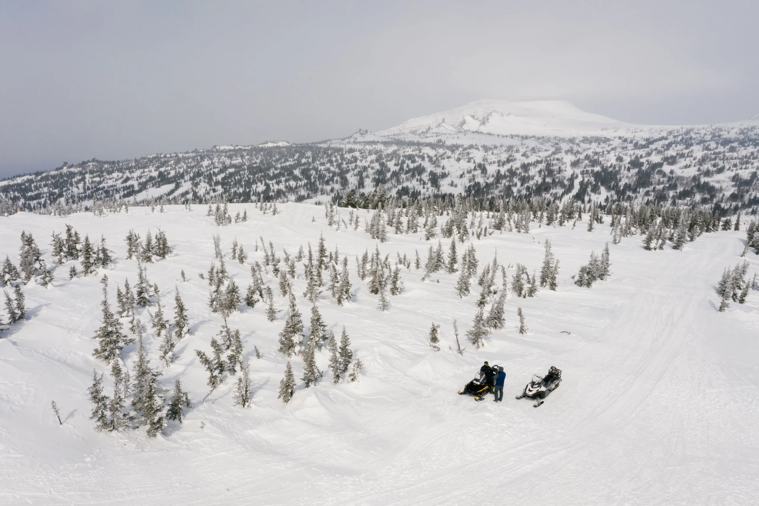Two small snowmobiles pause beside a winding trail in broad, snow-covered high country.