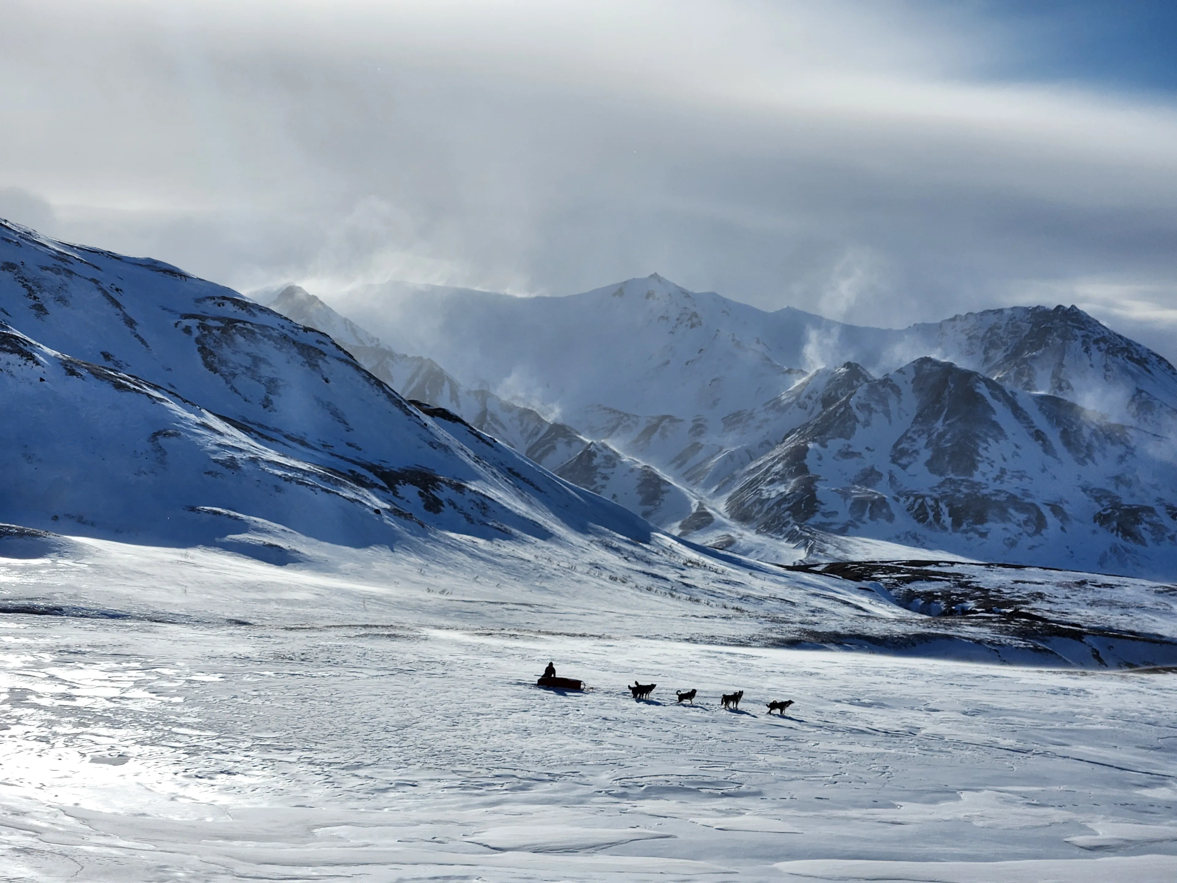A small dog team crosses an open snow basin below a vast range of windblown white mountains.