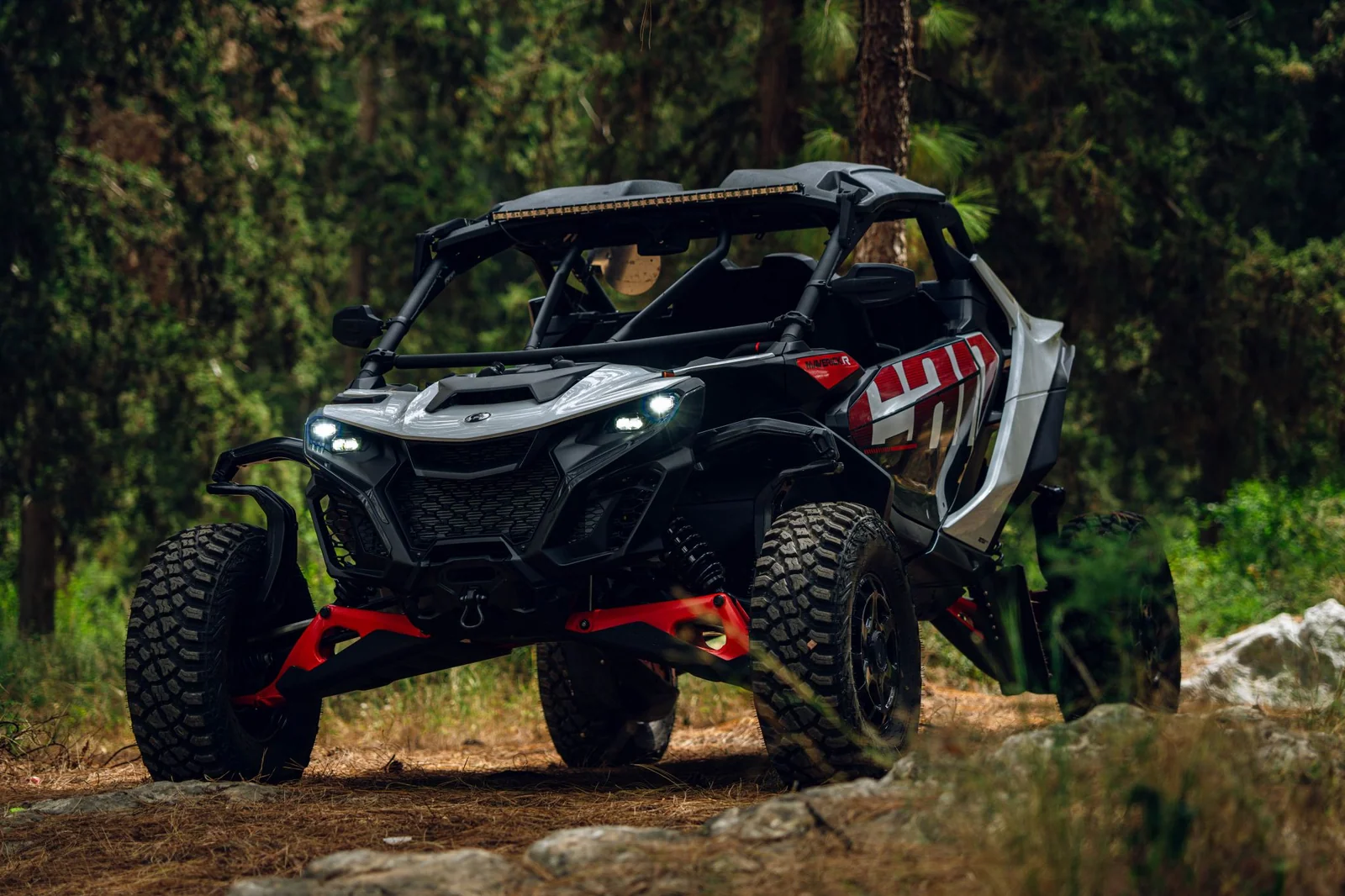 A two-seat Can-Am Maverick R parked on a woodland trail between evergreen trees.