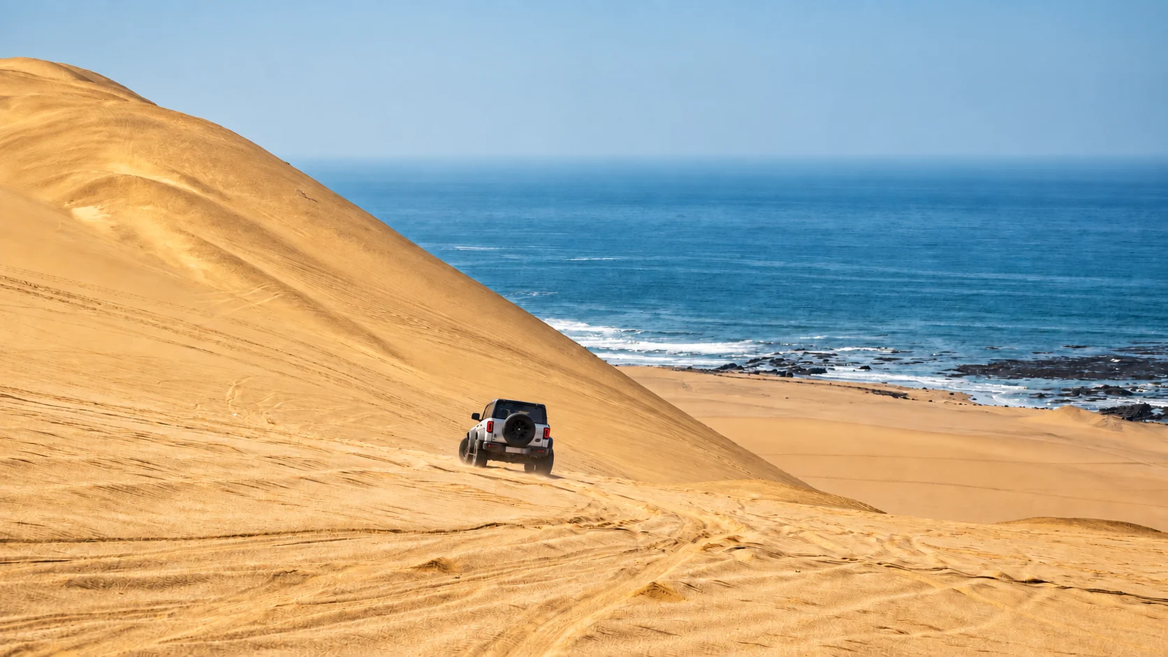 A white off-road vehicle descends a golden dune toward the ocean.