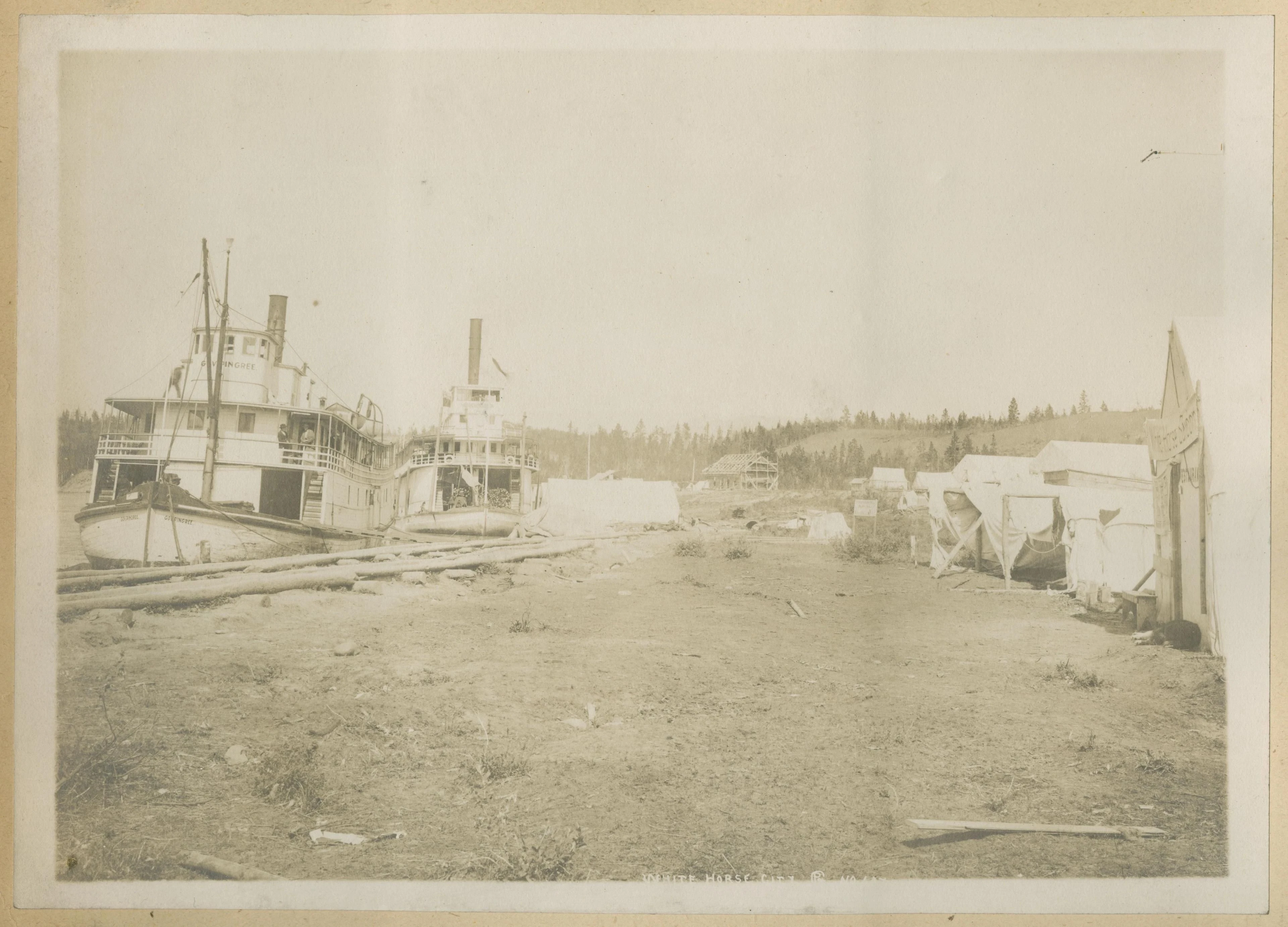 Two sternwheel riverboats beside a row of tents at Whitehorse in a pale sepia archival photograph.