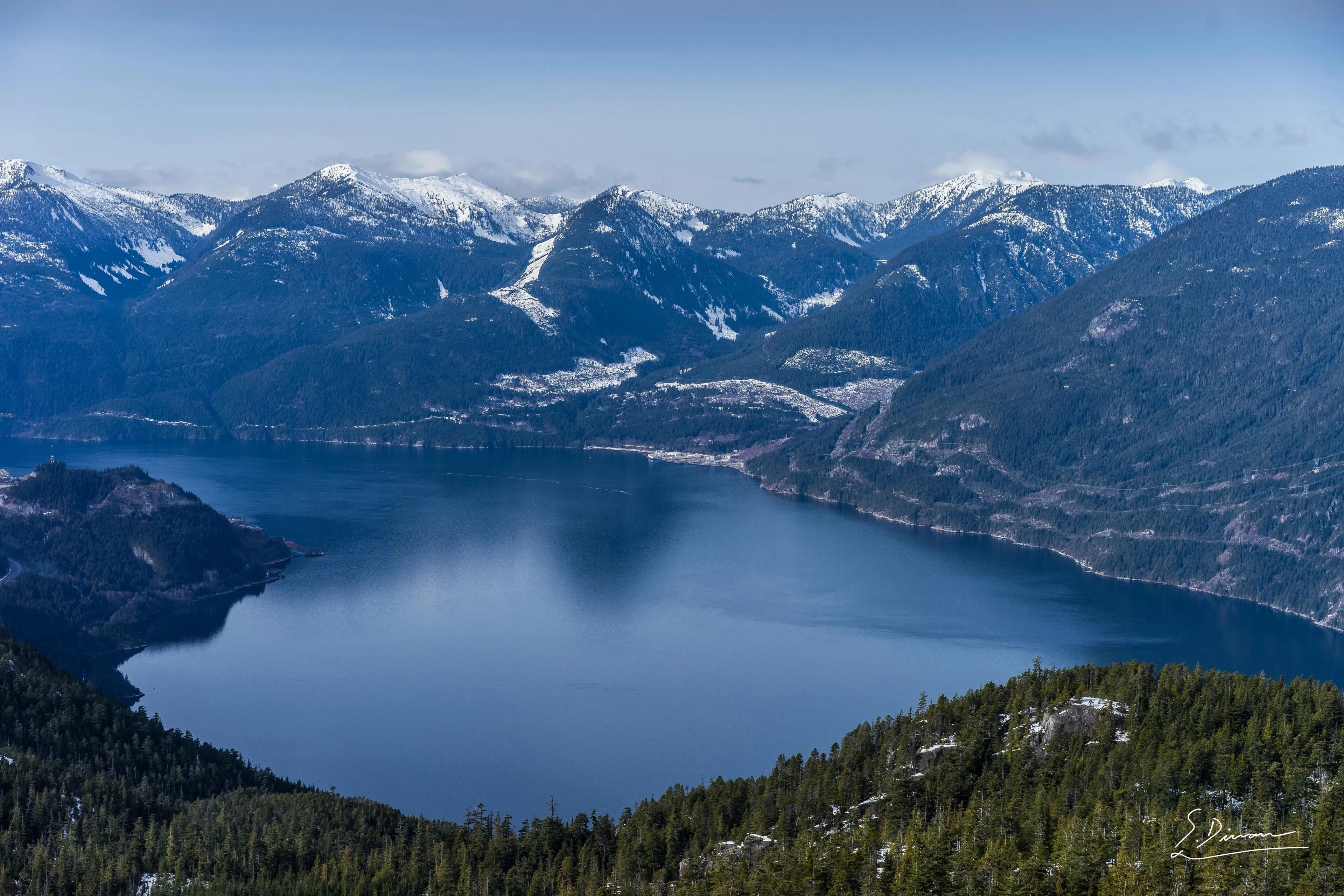 Blue water lies below forested slopes and snow-dusted mountains.