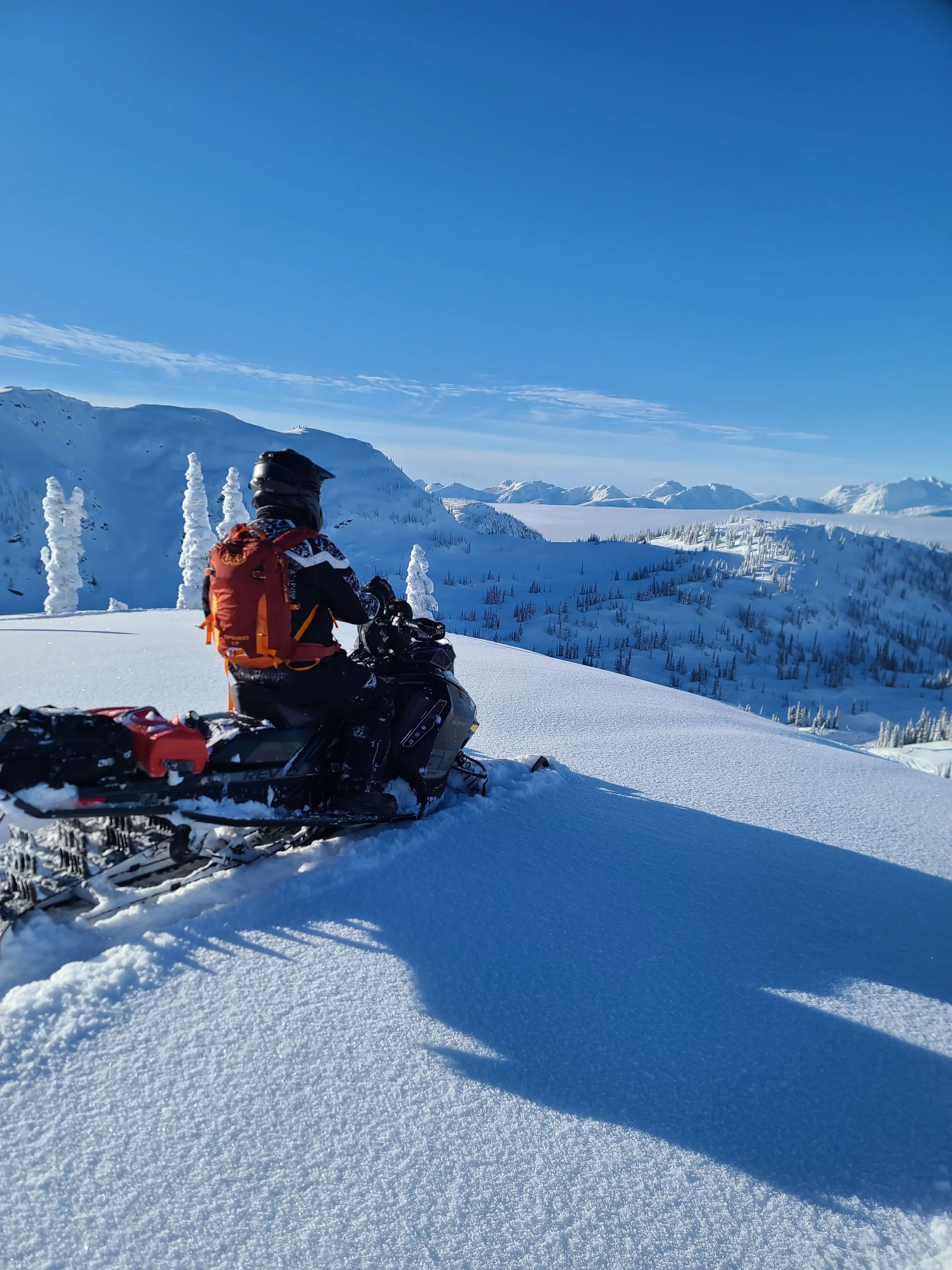 A snowmobile rider pauses on a snowy slope overlooking a sunlit mountain basin.