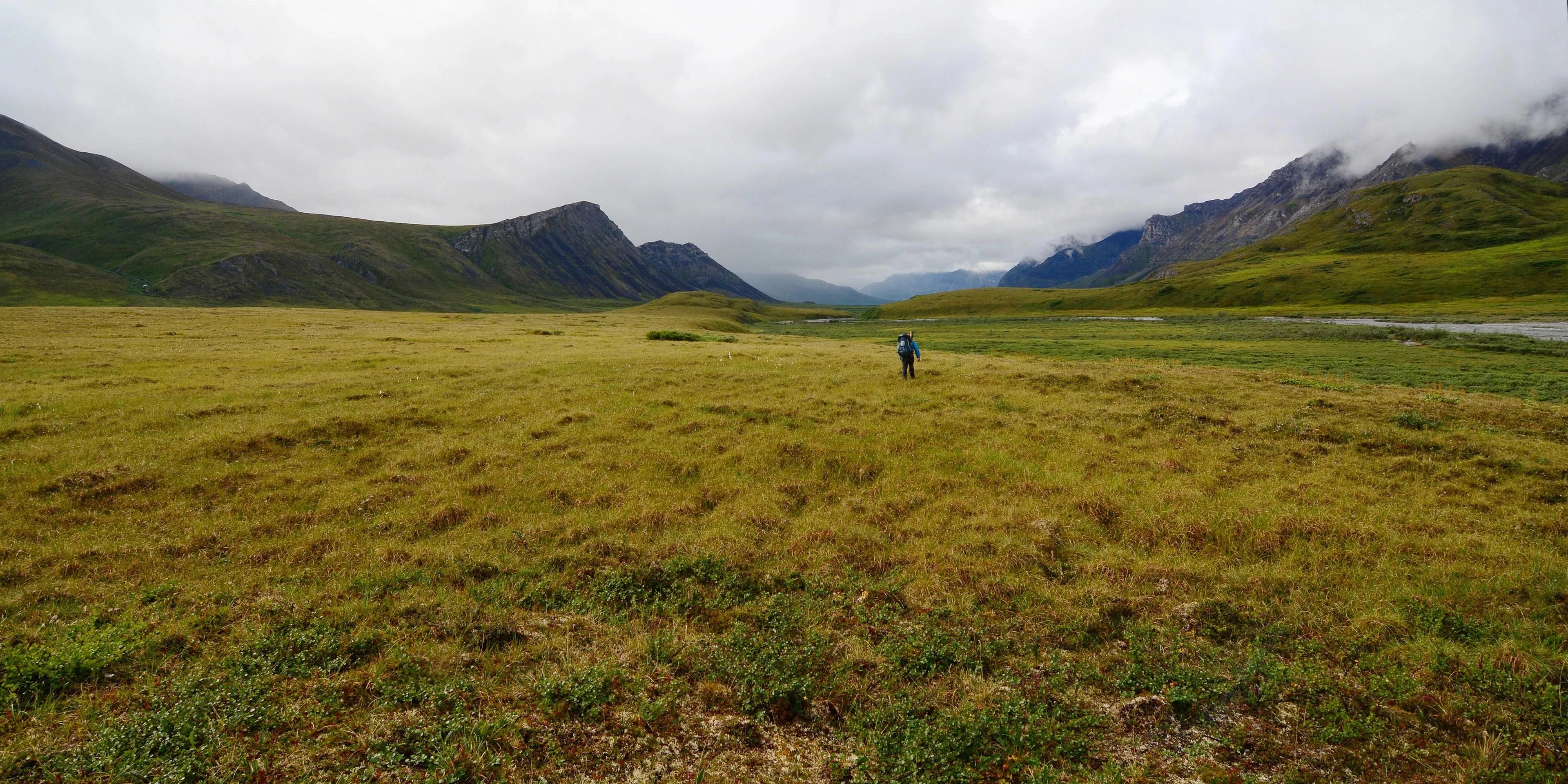 A small backpacker walks across a broad tundra valley beneath cloud-covered mountain slopes.