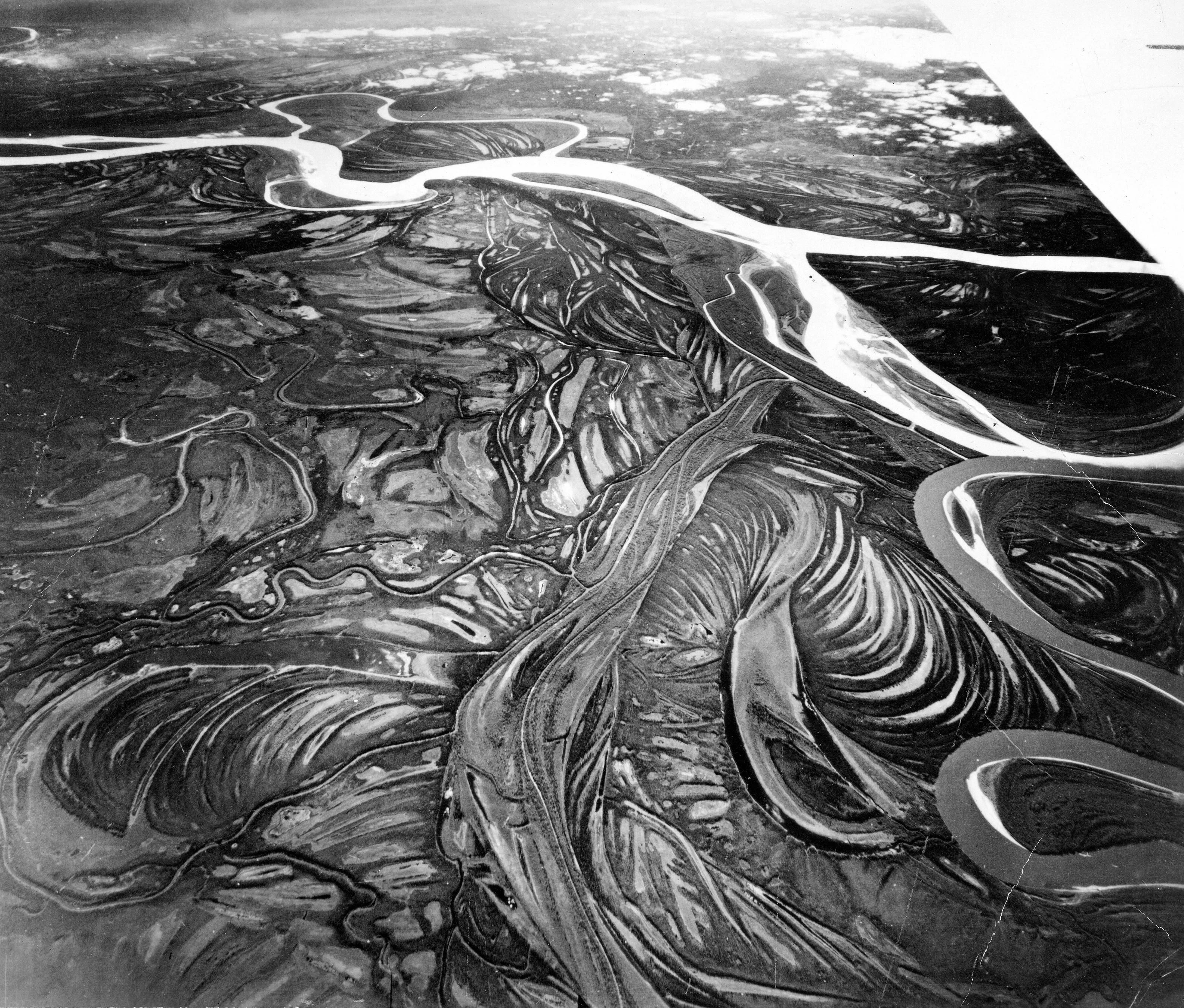 A black-and-white aerial photograph shows winding river channels and curved floodplain traces, with an aircraft wing at upper right.